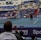 A tennis player stands on an indoor court holding a racket, ready to serve. The match is being watched by a sizeable audience, seated in blue and red stands. The umpire is visible on an elevated chair near the net. The court is well-lit, and various sponsors’ banners are displayed around the court edges.