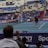 A tennis player stands on an indoor court holding a racket, ready to serve. The match is being watched by a sizeable audience, seated in blue and red stands. The umpire is visible on an elevated chair near the net. The court is well-lit, and various sponsors’ banners are displayed around the court edges.