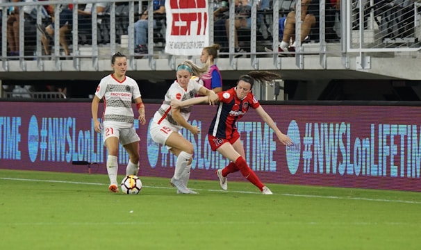 A competitive soccer match is underway with players vying for the ball near the sideline. The players wear different uniforms, indicating they are from opposing teams. The audience is visible in the stadium seating above, and there are visible advertisements and hashtags on the LED boards along the field.