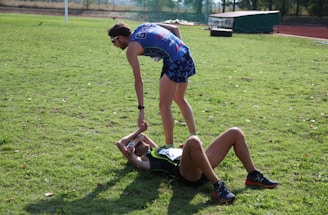 Two athletes are on a grassy field. One athlete is standing and offering a hand to another athlete who is lying on the ground. Both are wearing athletic gear, and it appears to be a moment of sportsmanship or assistance.