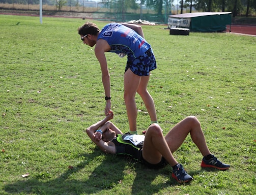 Two athletes are on a grassy field. One athlete is standing and offering a hand to another athlete who is lying on the ground. Both are wearing athletic gear, and it appears to be a moment of sportsmanship or assistance.