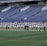 A group of athletes in matching white and dark-colored uniforms are standing in a line on a sports field. The background shows empty blue stadium seats and a wall with names inscribed on it.