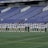 A group of athletes in matching white and dark-colored uniforms are standing in a line on a sports field. The background shows empty blue stadium seats and a wall with names inscribed on it.
