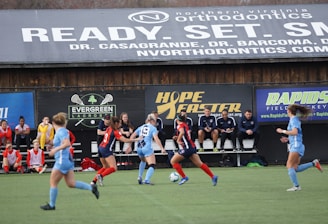 Players engage in a competitive soccer game on a field while others are seated on the bench watching attentively. The action is taking place in front of a wooden structure displaying multiple advertisements, including orthodontics, lacrosse, and field hockey.