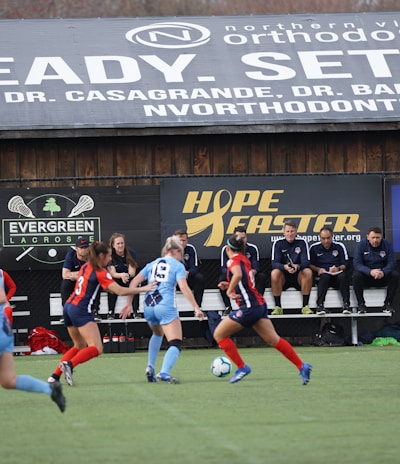 Players engage in a competitive soccer game on a field while others are seated on the bench watching attentively. The action is taking place in front of a wooden structure displaying multiple advertisements, including orthodontics, lacrosse, and field hockey.