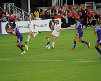 A soccer match in progress with players actively competing for the ball on a well-maintained field. The players are wearing team uniforms, and there is a crowd in the grandstand watching the game intently with some banners and advertisements visible in the background.