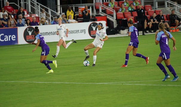 A soccer match in progress with players actively competing for the ball on a well-maintained field. The players are wearing team uniforms, and there is a crowd in the grandstand watching the game intently with some banners and advertisements visible in the background.