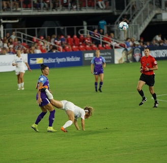 A soccer match is taking place on a well-maintained field. A player in a white jersey is falling forward, possibly after a physical interaction with a player in a purple jersey. Other players and a referee watch the event unfold. The stands are filled with spectators, and advertisements are visible along the sidelines.