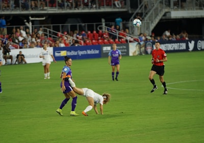 A soccer match is taking place on a well-maintained field. A player in a white jersey is falling forward, possibly after a physical interaction with a player in a purple jersey. Other players and a referee watch the event unfold. The stands are filled with spectators, and advertisements are visible along the sidelines.
