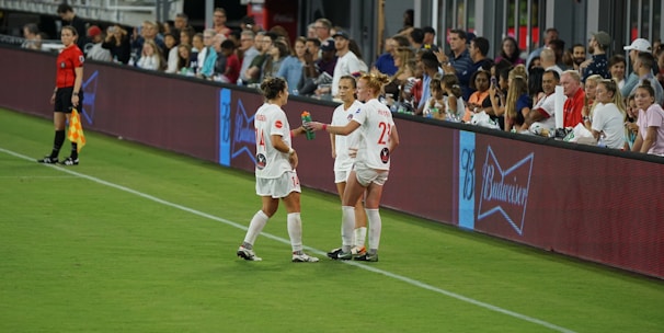 Two soccer players in white uniforms stand on the sideline of a field near a digital advertising board. One player holds a beverage bottle, and they appear to be in conversation. A referee in black stands nearby, and spectators fill the stands in the background, watching the event.