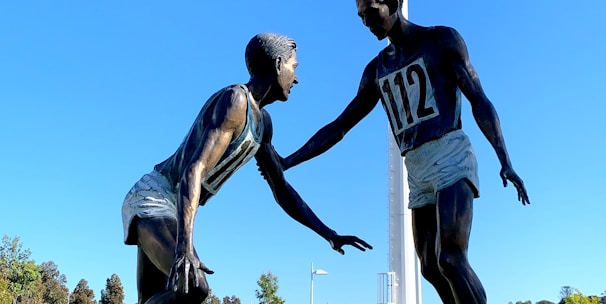 A sculpture depicting two athletes in mid-action is prominently displayed against a clear blue sky. One figure is reaching out to help the other, illustrating a moment of sportsmanship. The bronze statues stand on a black pedestal with an inscription, situated in an open green field with trees and sports facilities in the background. A tall floodlight tower looms overhead, adding to the outdoor sports setting.