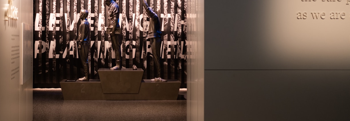 A dimly lit display features statues of athletes standing on a podium. They are positioned in front of a backdrop with the words 'LEVELING THE PLAYING FIELD' in bold letters. On the wall to the right, a softly lit quote is partially visible. The overall ambiance suggests a museum or exhibition setting.