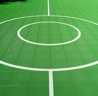A vibrant green sports court featuring distinct white circular markings and straight lines. The court surface is composed of a textured grid pattern, and small leaves can be seen scattered on the surface.