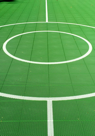 A vibrant green sports court featuring distinct white circular markings and straight lines. The court surface is composed of a textured grid pattern, and small leaves can be seen scattered on the surface.