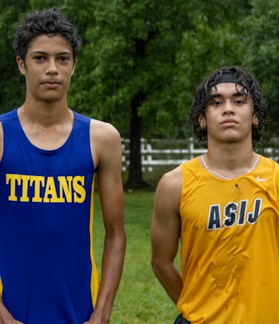 Two young athletes wearing sleeveless sports tops stand on a grassy field. The athlete on the left is wearing a blue top with the word 'TITANS' while the one on the right wears a yellow top with 'ASIJ'. The background features green trees and a white fence, with a bench visible on the right.