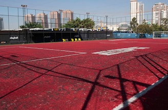 A sports court with a red playing surface and white lines, surrounded by a black mesh fence. In the background, tall buildings are visible, along with some trees and a few yellow chairs positioned near the fence.
