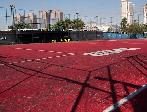 A sports court with a red playing surface and white lines, surrounded by a black mesh fence. In the background, tall buildings are visible, along with some trees and a few yellow chairs positioned near the fence.